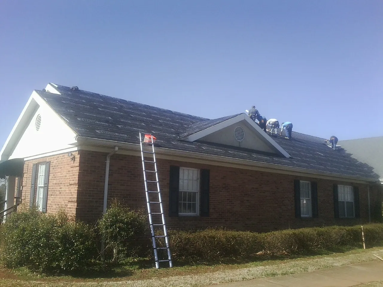 Crew tearing shingles off a brick ranch roof with a ladder set — Emergency Roof Repair work by our crew in Gastonia