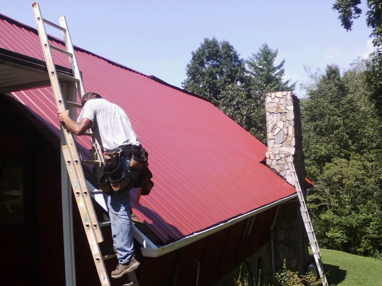 Roofer on a ladder beside a red metal roof and stone chimney — Soffit & Fascia Repair by Roofing Brothers in Gastonia